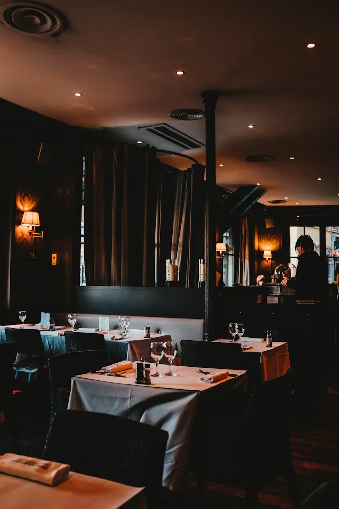 A dimly lit dining room with white-clothed tables and warm wall lights