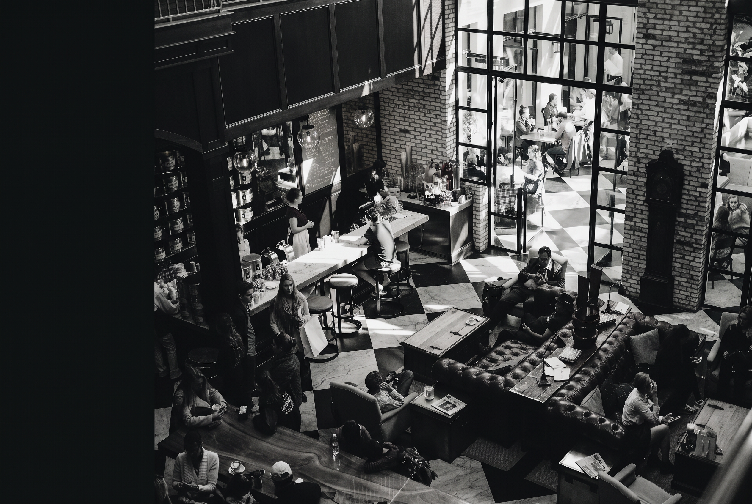 Black-and-white photograph of a bustling café and bar interior with guests at tables and a leather sofa